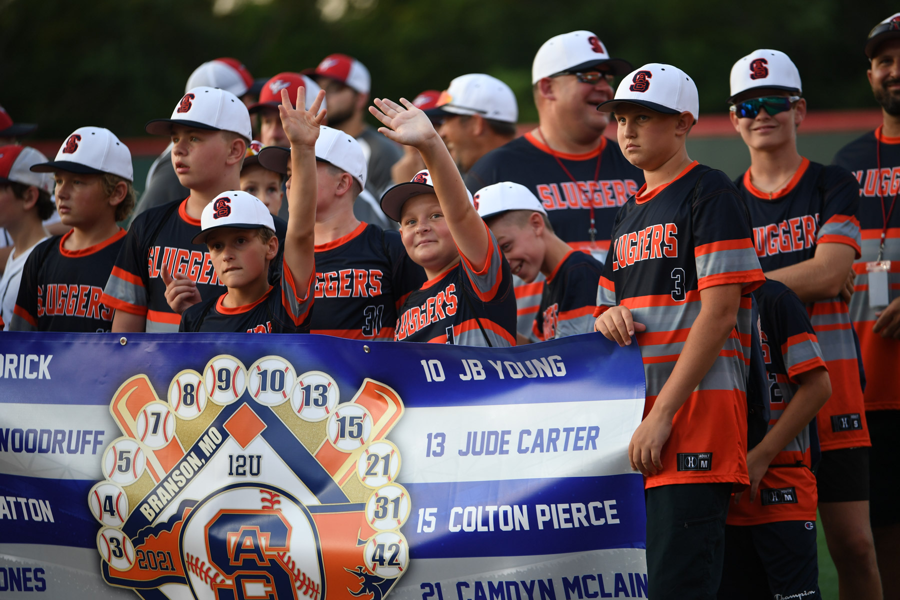 A youth baseball team in black and orange uniforms stand together holding a banner in front of them.