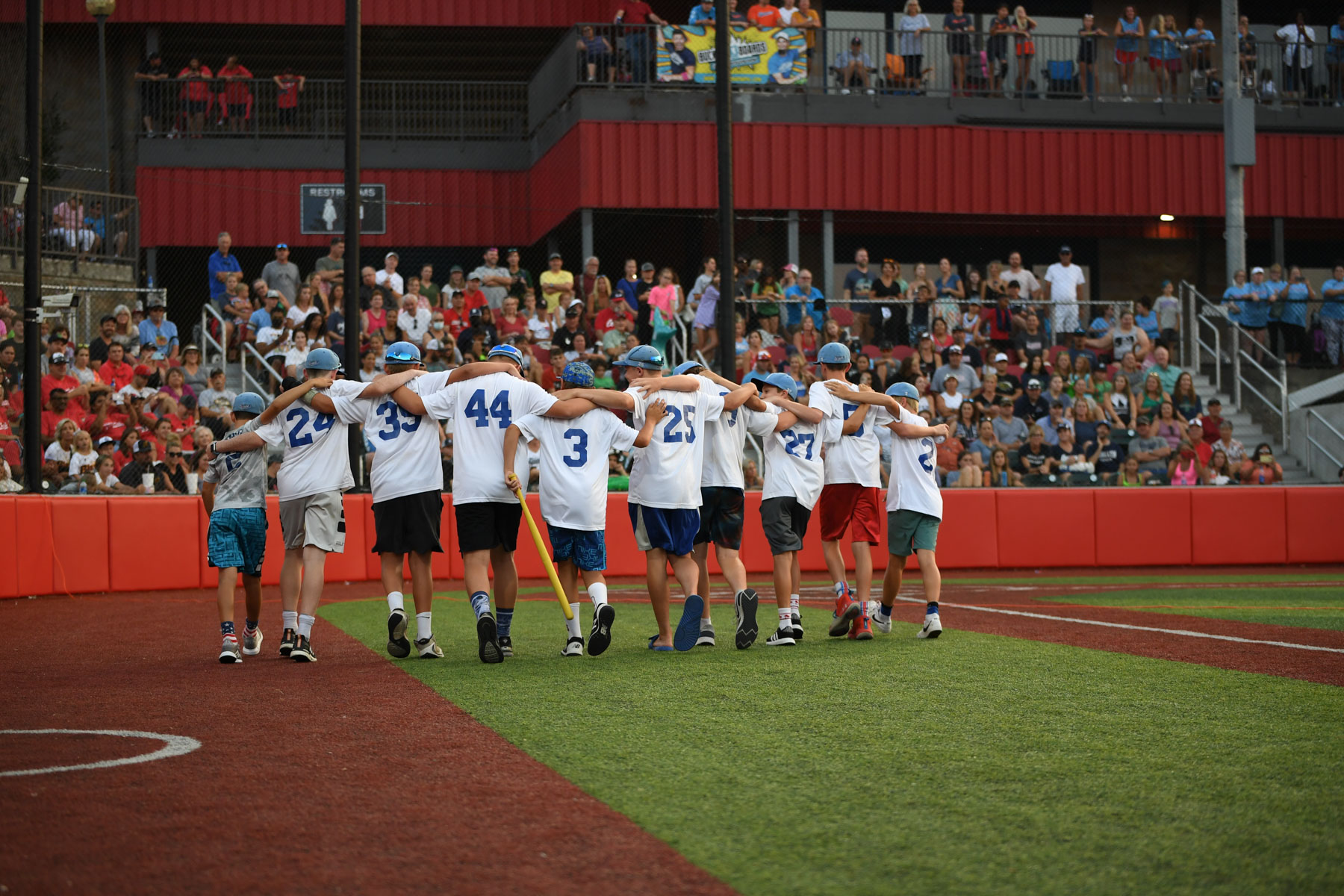 A team of youth travel baseball players have their arms around each other's shoulders as they walk off a baseball field. The players are wearing white jerseys with blue numbers.